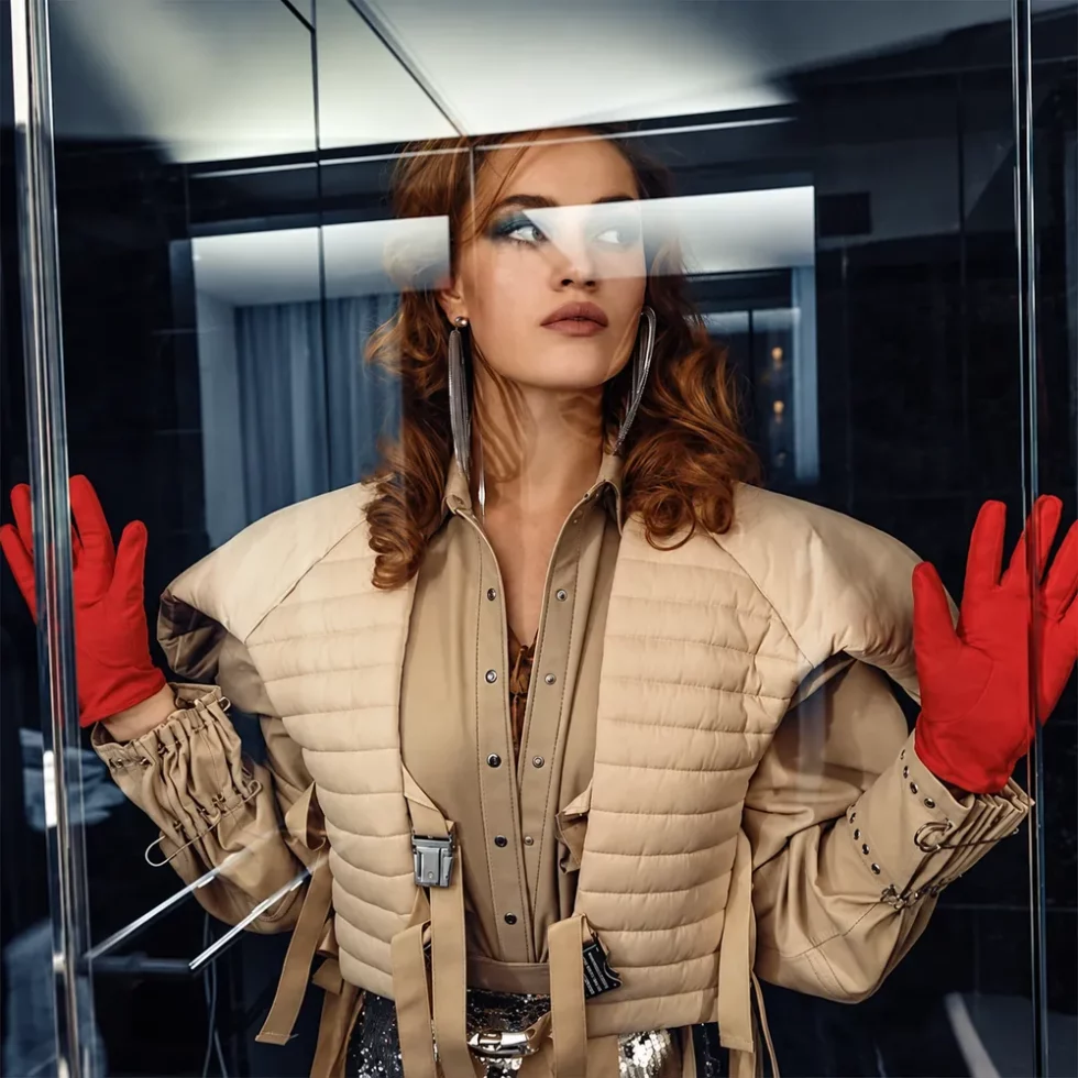 Fashion-Editorial: Model in beiger Steppjacke mit roten Handschuhen hinter einer Glasscheibe